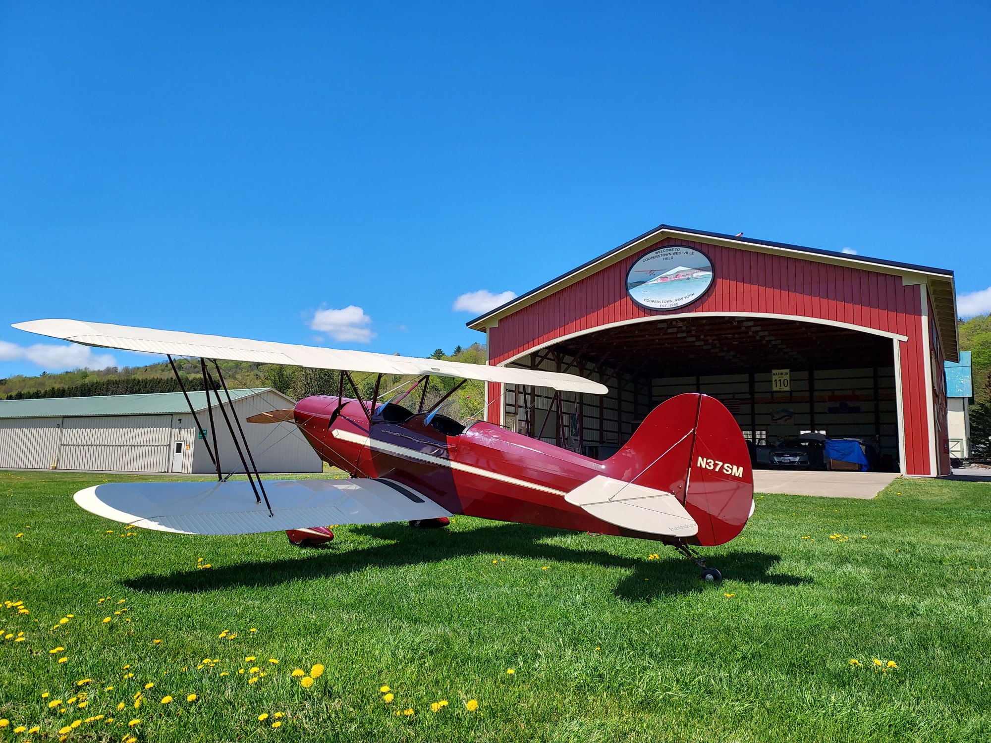 Red biplane parked on the grass with hangar behind