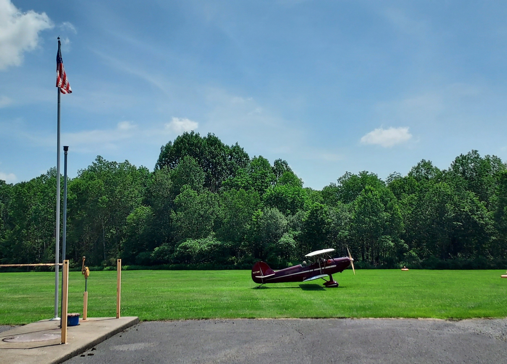 Burgundy biplane parked on the grass airfield with American flag and tree line